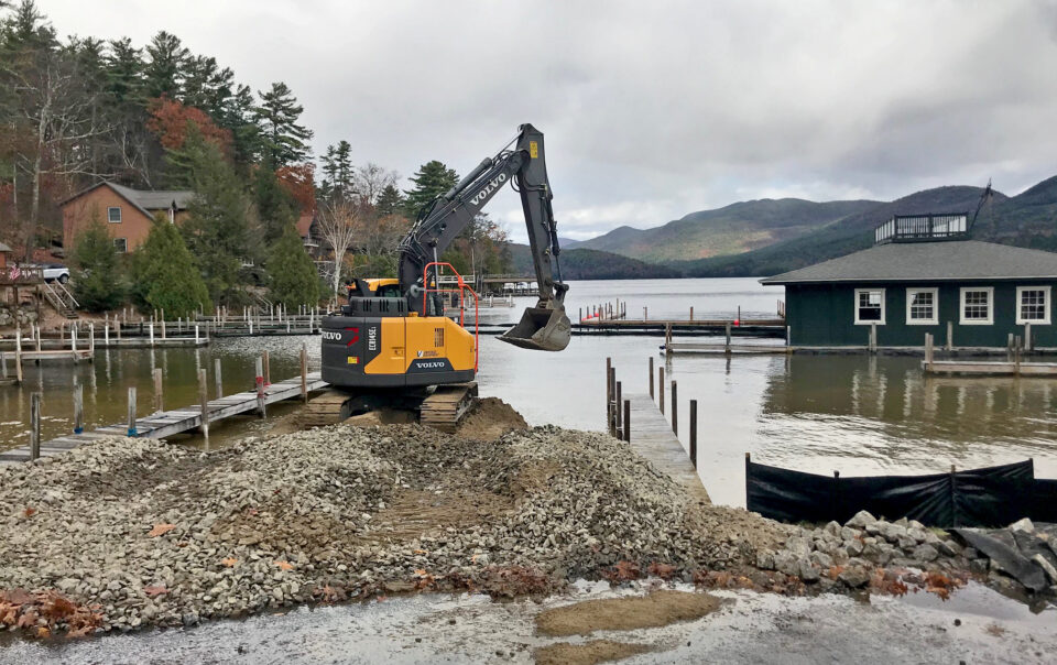 Excavator moving gravel on waterfront site design project with lake and mountain landscape in Northeast region.