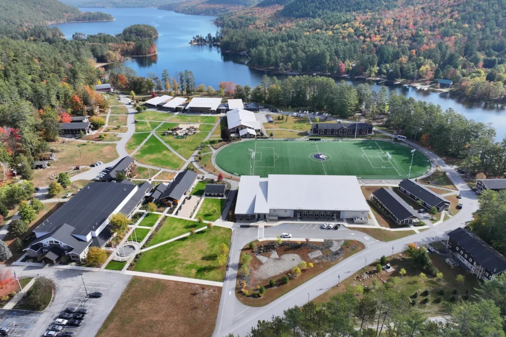 Aerial view of waterfront campus with sports field, buildings, parking, and forested landscape overlooking blue lake