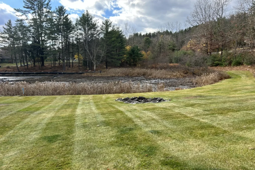 Waterfront landscape with manicured lawn, fire pit, native plantings, and mature trees overlooking pond in Northeast setting