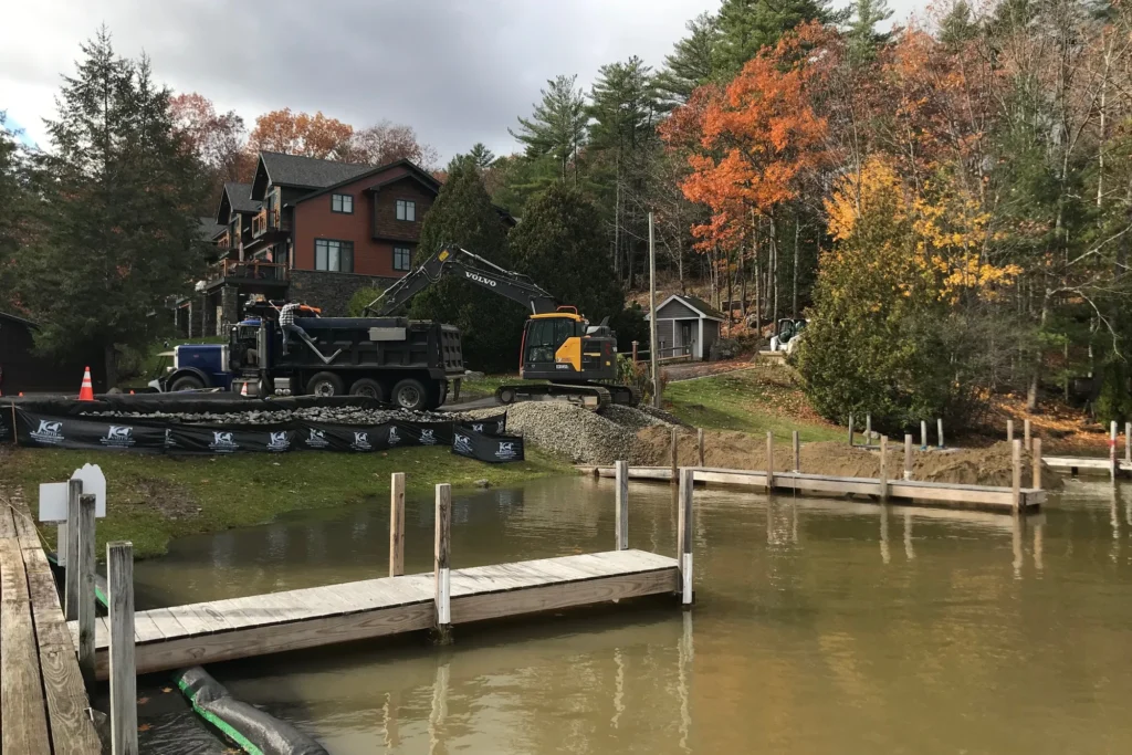 Waterfront property development with dock, excavator, dump truck, and residential home during fall season in Northeast