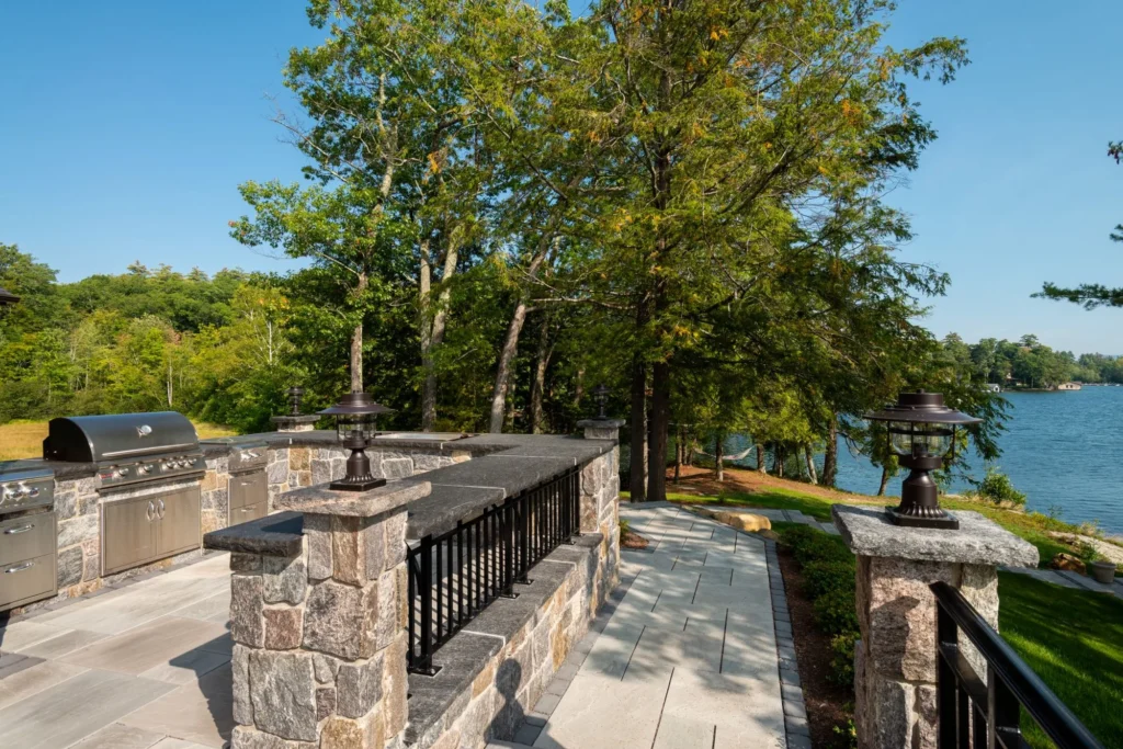 Waterfront outdoor kitchen and patio with stone construction, mature trees, and lakeside seating area for entertaining.