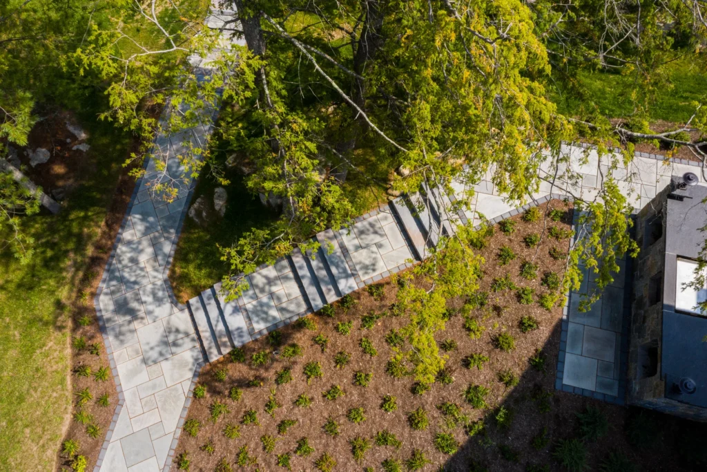 Aerial view of modern landscape design with paved walkway, planted beds, and mature trees framing the site.