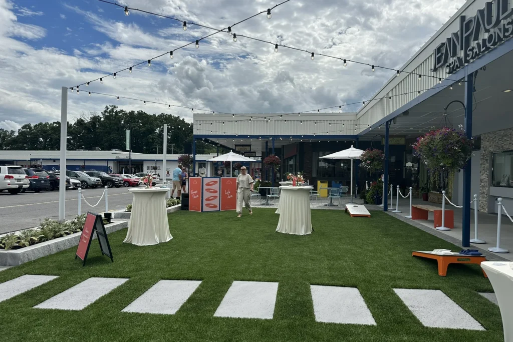 Outdoor event space with striped pavement, lawn, string lights, and retail storefronts showcasing site design