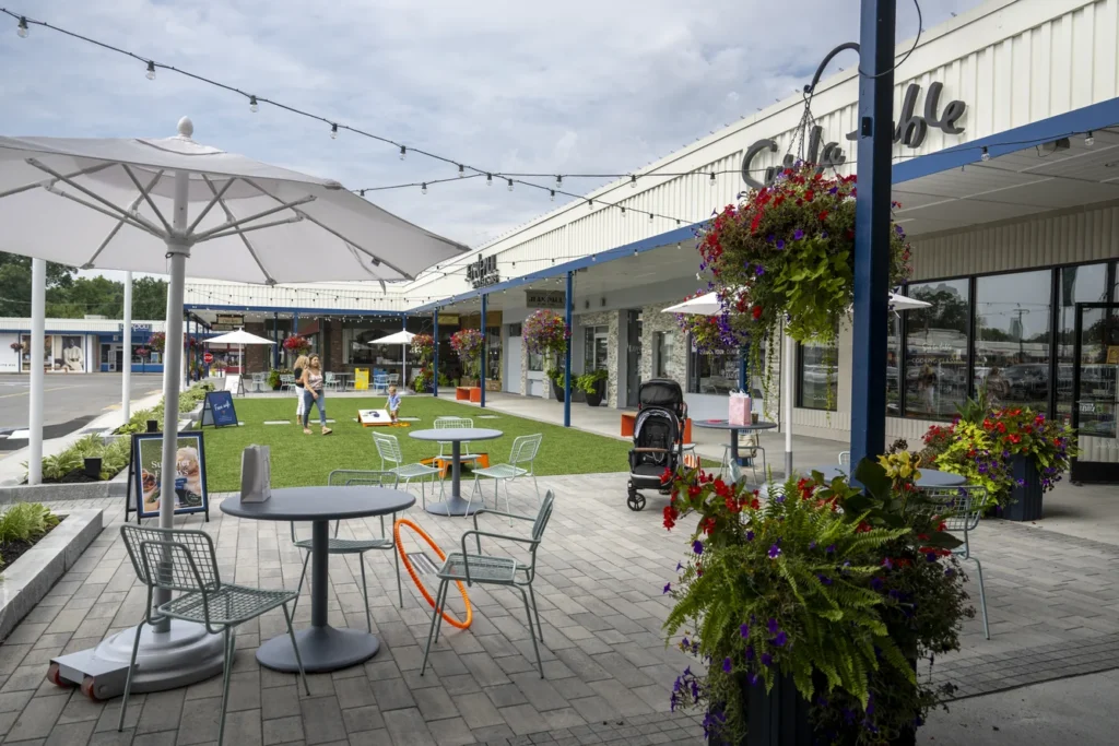 Modern plaza design with outdoor seating, vibrant hanging baskets, and mixed-use retail storefronts under shade structures.