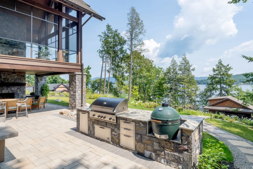 Waterfront patio with stone outdoor kitchen, grills, and lake views framed by tall trees