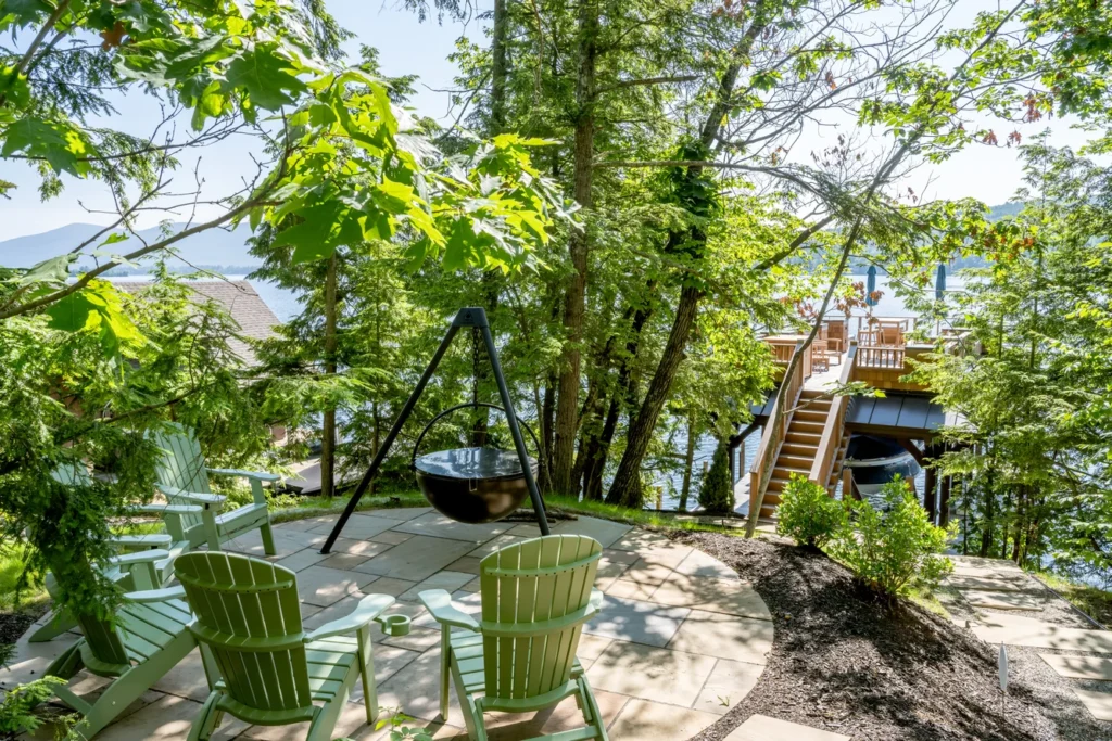 Waterfront patio with fire pit, Adirondack chairs, and mature trees overlooking lake and residential structures