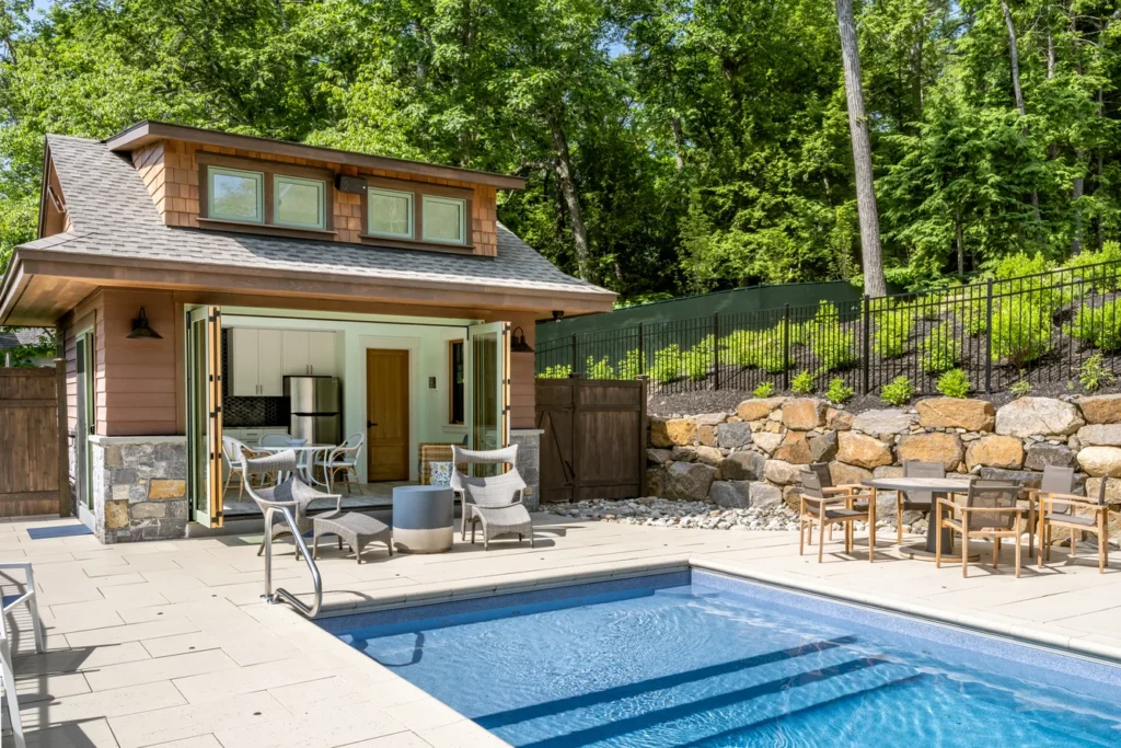 Modern residential pool area with stone retaining wall, patio, guest house, and landscaped grounds surrounded by mature