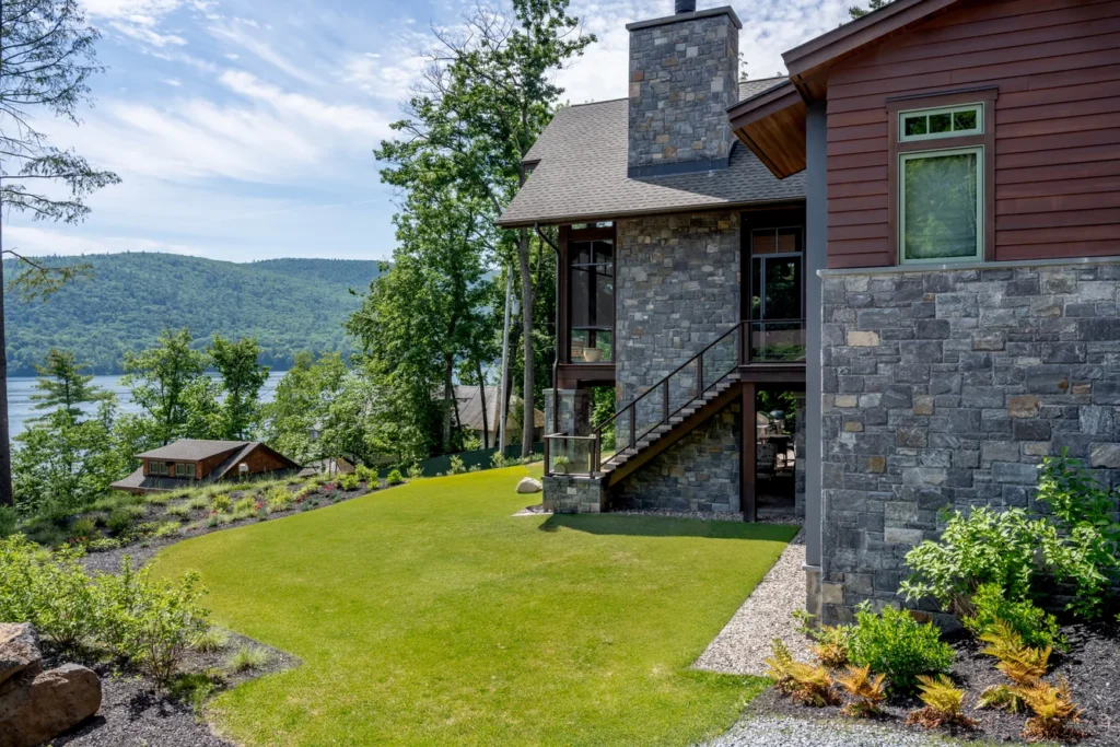 Modern waterfront home with manicured lawn, stone exterior, and native plantings overlooking forested hills and lake
