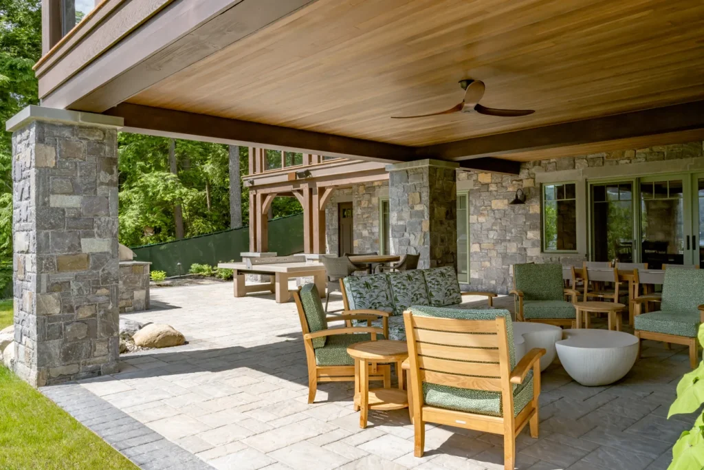 Covered patio with stone columns, wooden ceiling, and seating area overlooking landscaped grounds and green space.