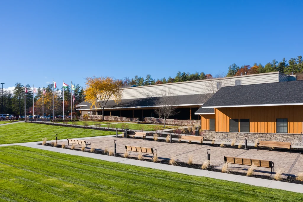 Modern civic building with stone and wood exterior, paved plaza with benches, landscaping, and fall foliage in Northeast